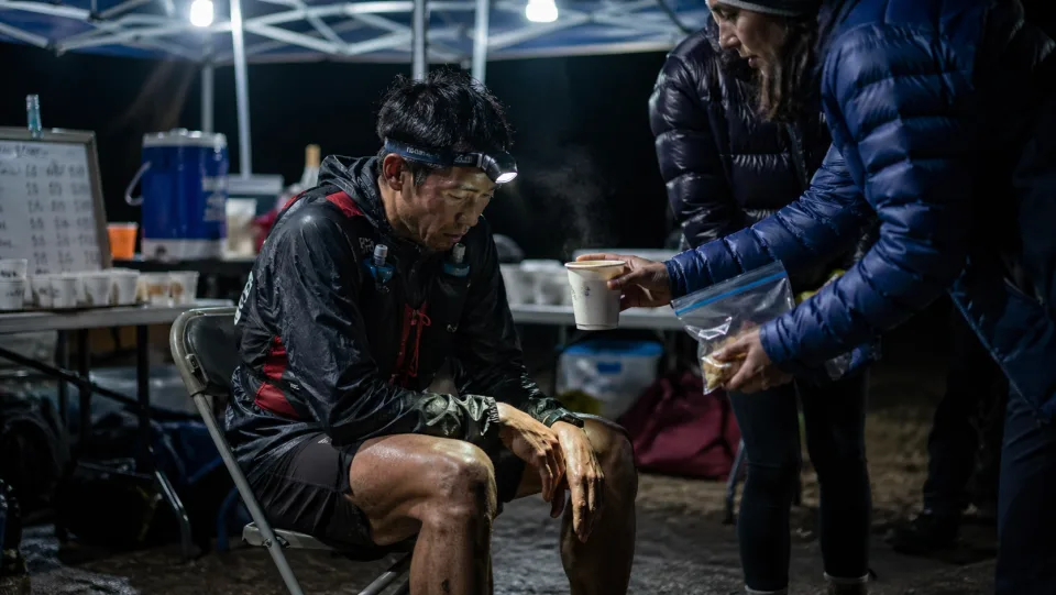 Ultrarunner refueling at a night aid station during an ultramarathon while support crew hands over warm food and drink