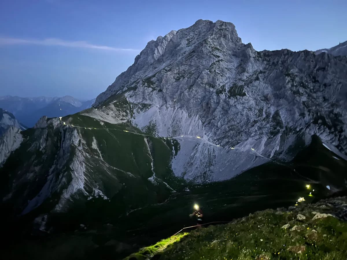 Headlamp lights of night runners below a massive limestone face at dusk